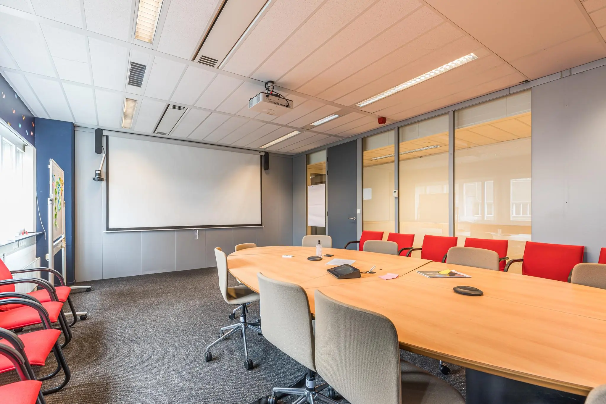 Vergaderzaal aan het Leidseveer met ovale tafel, rode en grijze stoelen, projectiescherm en glazen binnenwand.