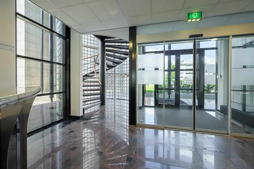 Ingang van een modern kantoor aan de Rivium Westlaan met glazen deuren, een wenteltrap en marmeren vloer.