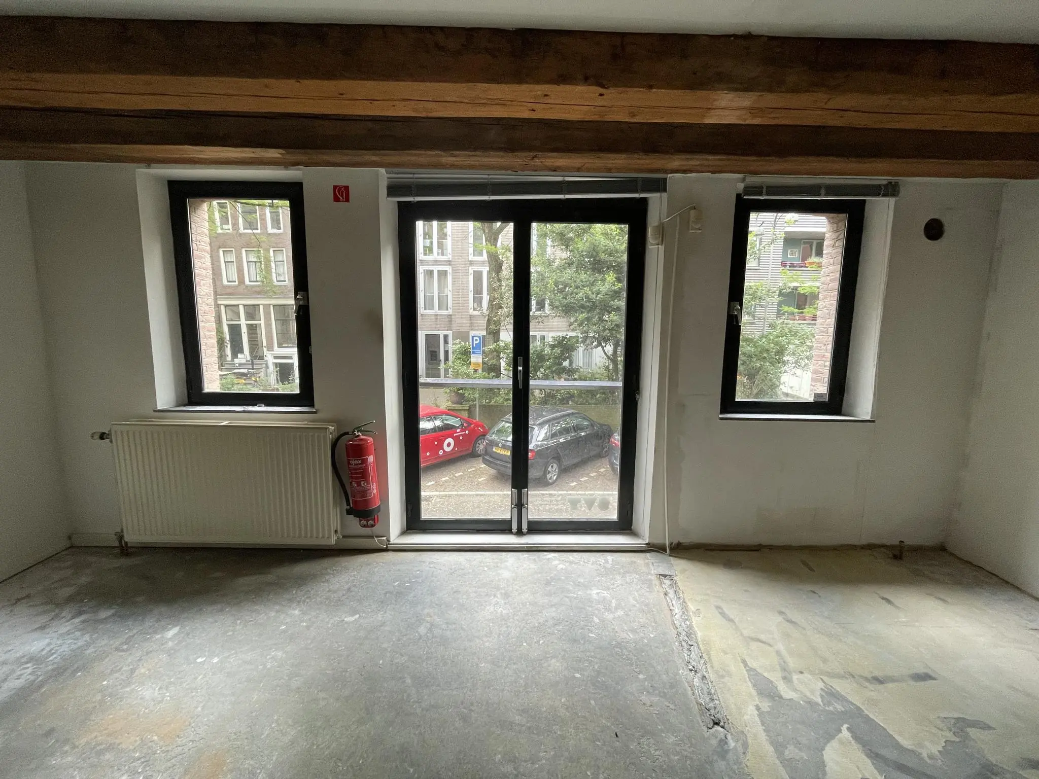 Uitzicht vanuit een lege kamer op het Entrepotdok met drie ramen, een brandblusser en zicht op geparkeerde auto's buiten.