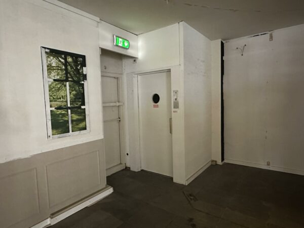 Een hoek van een kaal interieur met een witte deur, een nepraam met natuurafbeelding en een groen nooduitgangsbord aan het plafond.