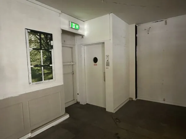 Een hoek van een kaal interieur met een witte deur, een nepraam met natuurafbeelding en een groen nooduitgangsbord aan het plafond.