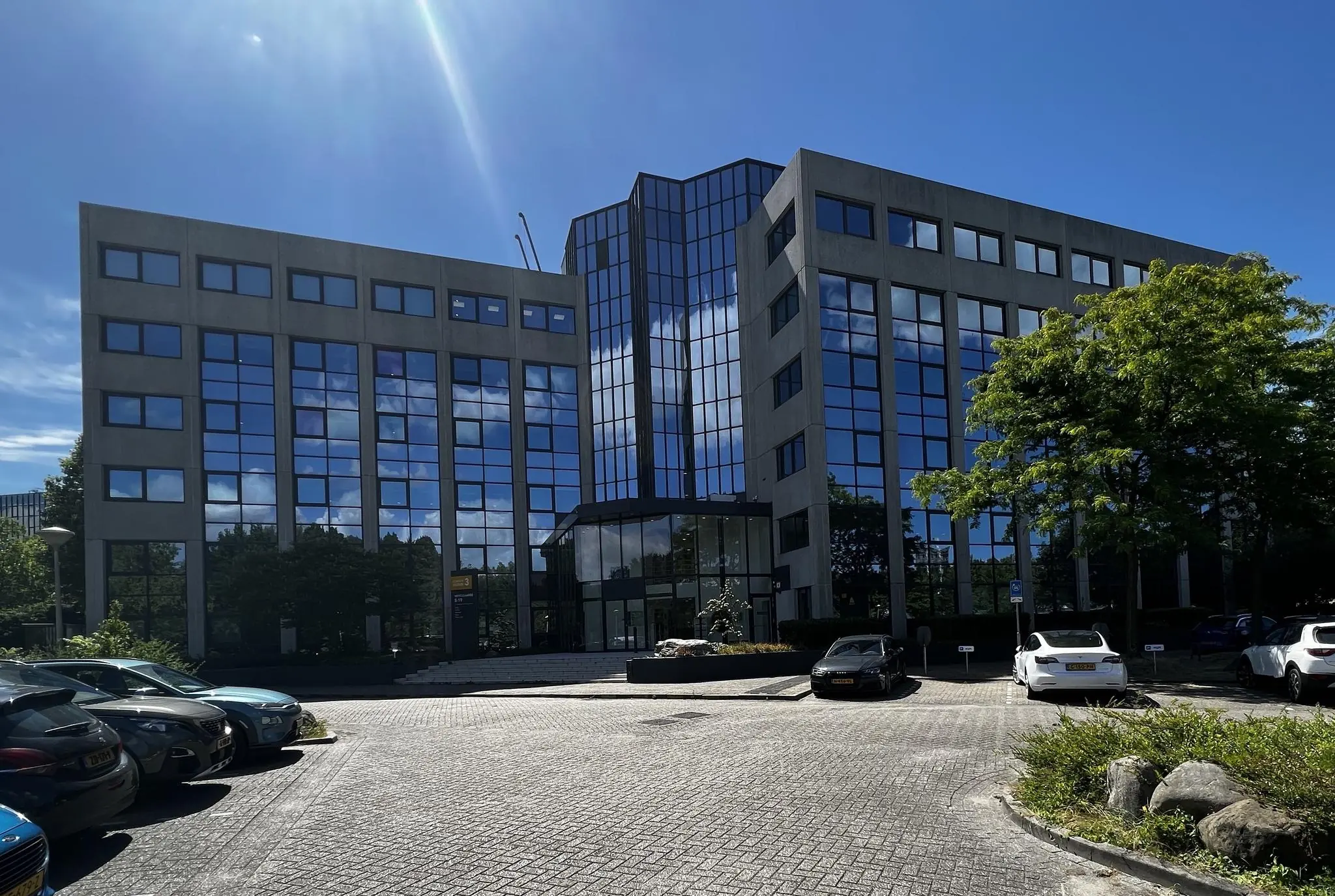 Kantoorgebouw aan de Nevelgaarde met spiegelende ramen en geparkeerde auto's op een zonnige dag.