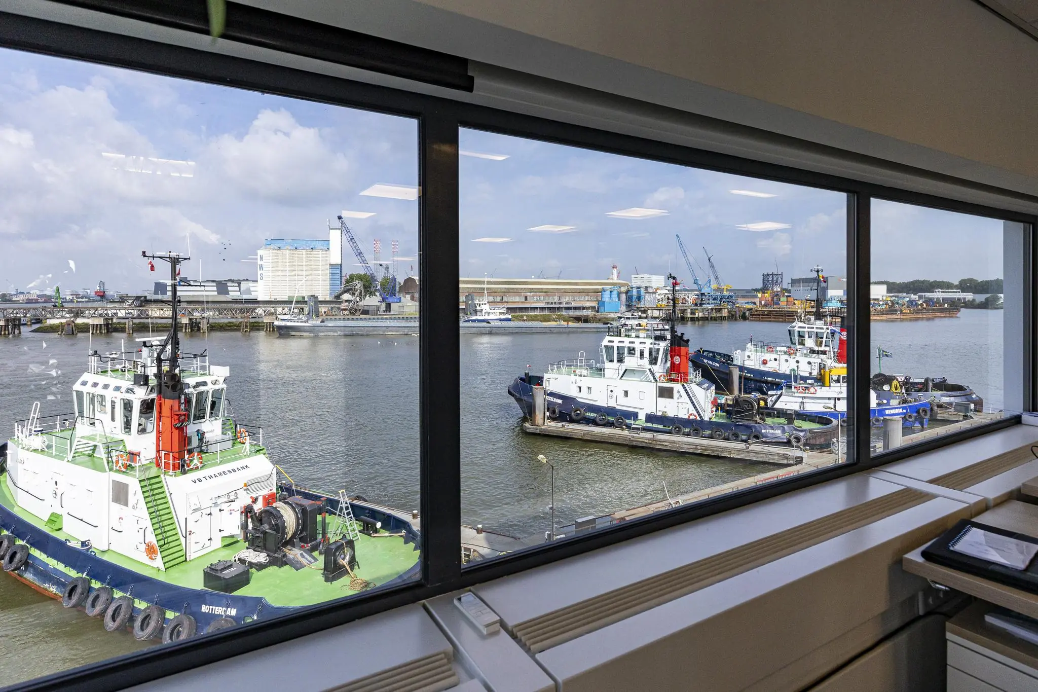 Uitzicht vanuit een kantoorraam op sleepboten aangemeerd aan de Havenstraat in een industriële havenomgeving.