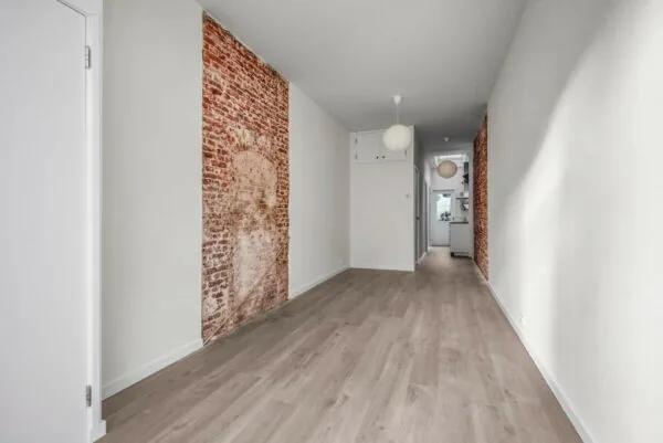 Langwerpige, smalle woonkamer met een houten vloer, witte muren en twee decoratieve bakstenen muurdelen aan weerszijden.