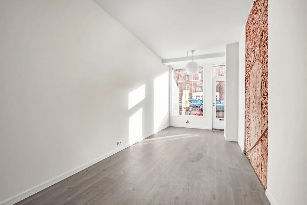 Lichte, lege kamer aan de Westerstraat met een houten vloer, grote ramen en een muur met bakstenen accent.