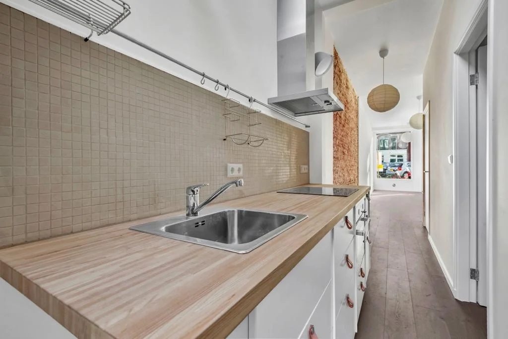 Moderne smalle keuken in een appartement aan de Westerstraat met lichte houten werkbladen en een beige mozaïektegel achterwand.