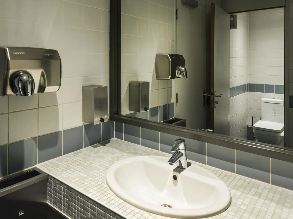 Wastafel met spiegel in een openbare toiletruimte, met zicht op een gesloten toiletcabine.