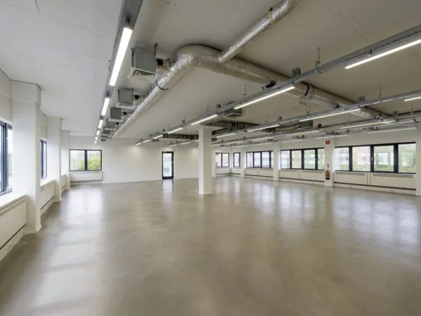 Lege, moderne kantoorruimte met betonnen vloer, witte muren, grote ramen en zichtbaar plafond met ventilatiebuizen.