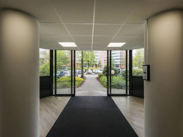 Ingang van een modern kantoorgebouw met grote glazen deuren en zicht op een parkeerplaats en kantoorpanden buiten.