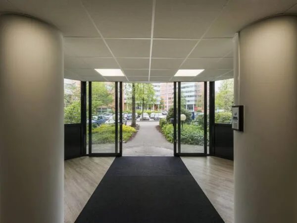 Ingang van een modern kantoorgebouw met grote glazen deuren en zicht op een parkeerplaats en kantoorpanden buiten.