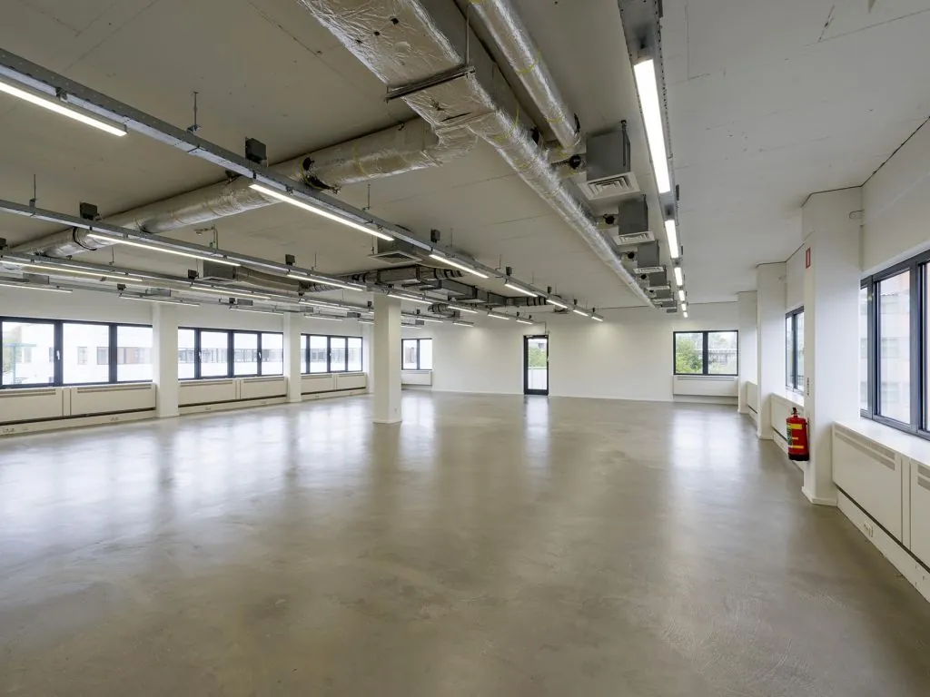 Leeg, modern kantoorpand met betonnen vloer, zichtbare plafondinstallaties en grote ramen voor natuurlijk licht.