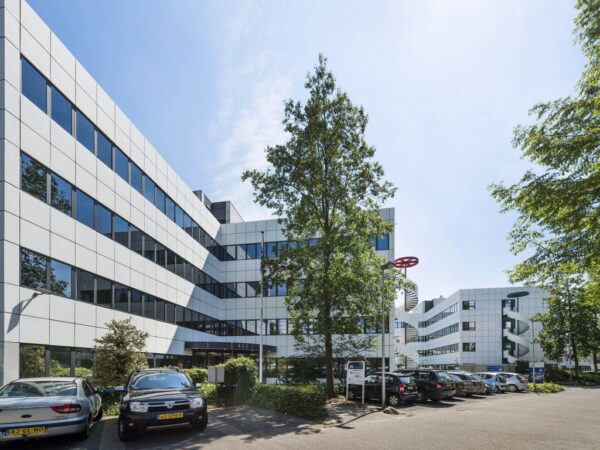 Wit kantoorgebouw met spiegelende ramen en een parkeerplaats op de voorgrond.