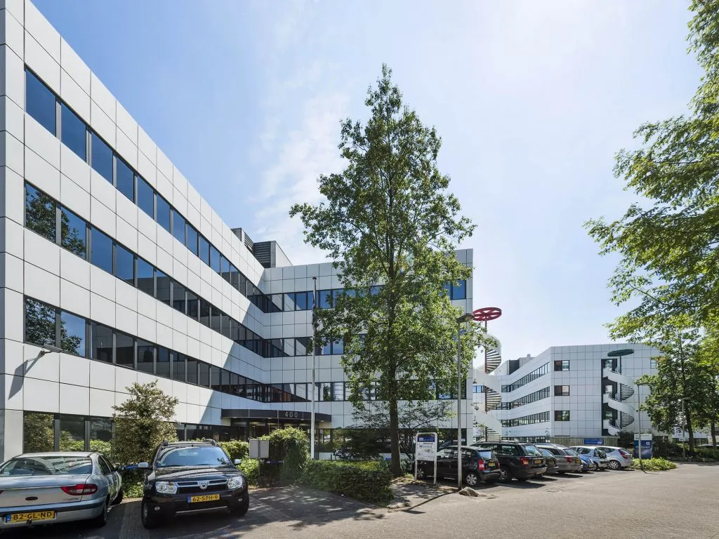 Wit kantoorgebouw met spiegelende ramen en een parkeerplaats op de voorgrond.