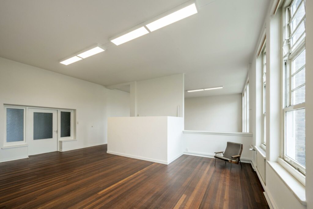 Lichte, minimalistische ruimte aan het WG-plein met houten vloer, grote ramen en een enkele stoel.