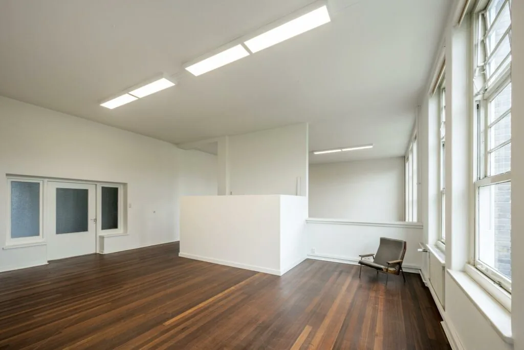 Lichte, minimalistische ruimte aan het WG-plein met houten vloer, grote ramen en een enkele stoel.