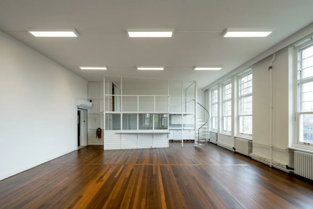 Lichte ruimte met houten vloer, grote ramen en een witte spiltrap naar een verhoogde werkplek aan het WG-plein.