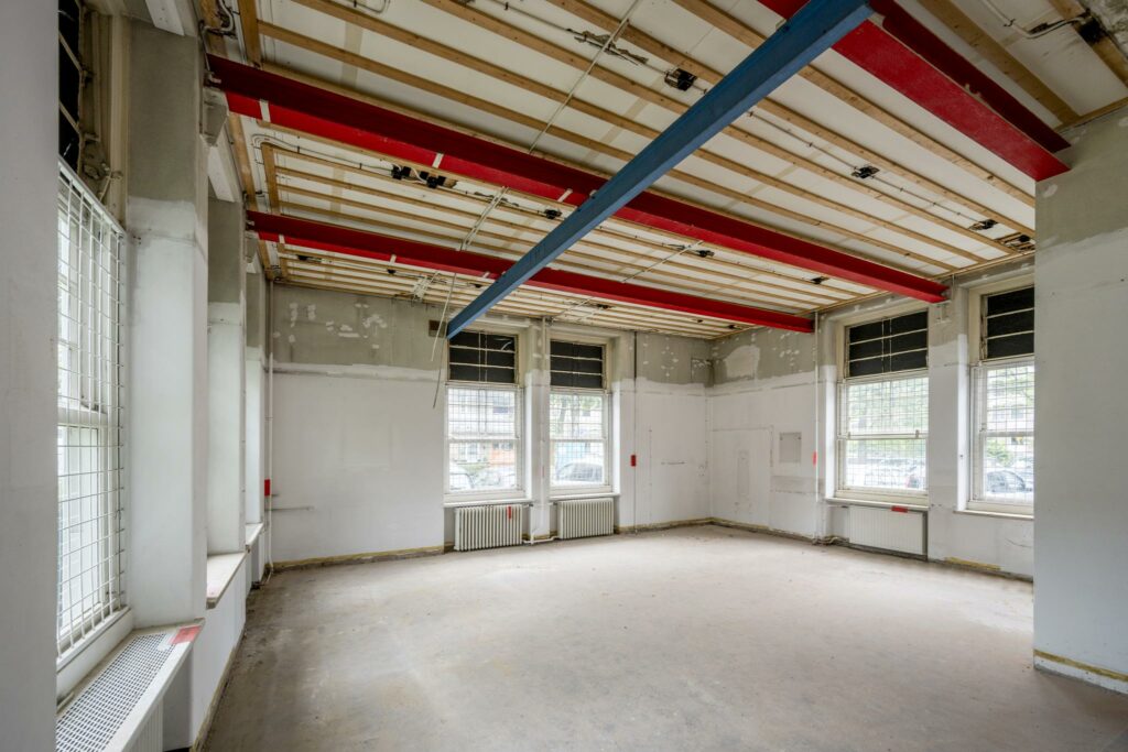 Lege, deels gerenoveerde ruimte met zichtbare plafondbalken en grote ramen aan het WG-plein.