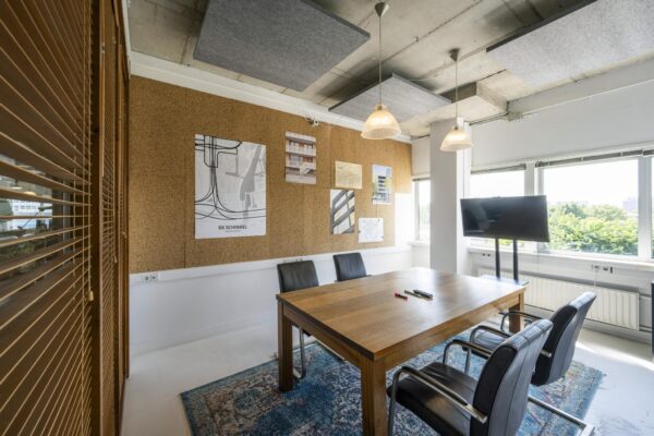 Vergaderruimte met houten tafel, vier stoelen, een prikbord met projectposters en een tv-scherm bij het raam aan de Anthony Fokkerweg.