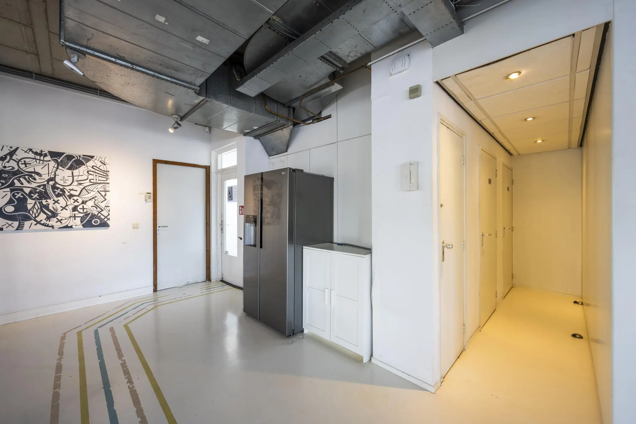 Witte gang met meerdere deuren, een grote grijze koelkast, zichtbare ventilatiebuizen en een abstract schilderij aan de muur in een pand aan de Anthony Fokkerweg.