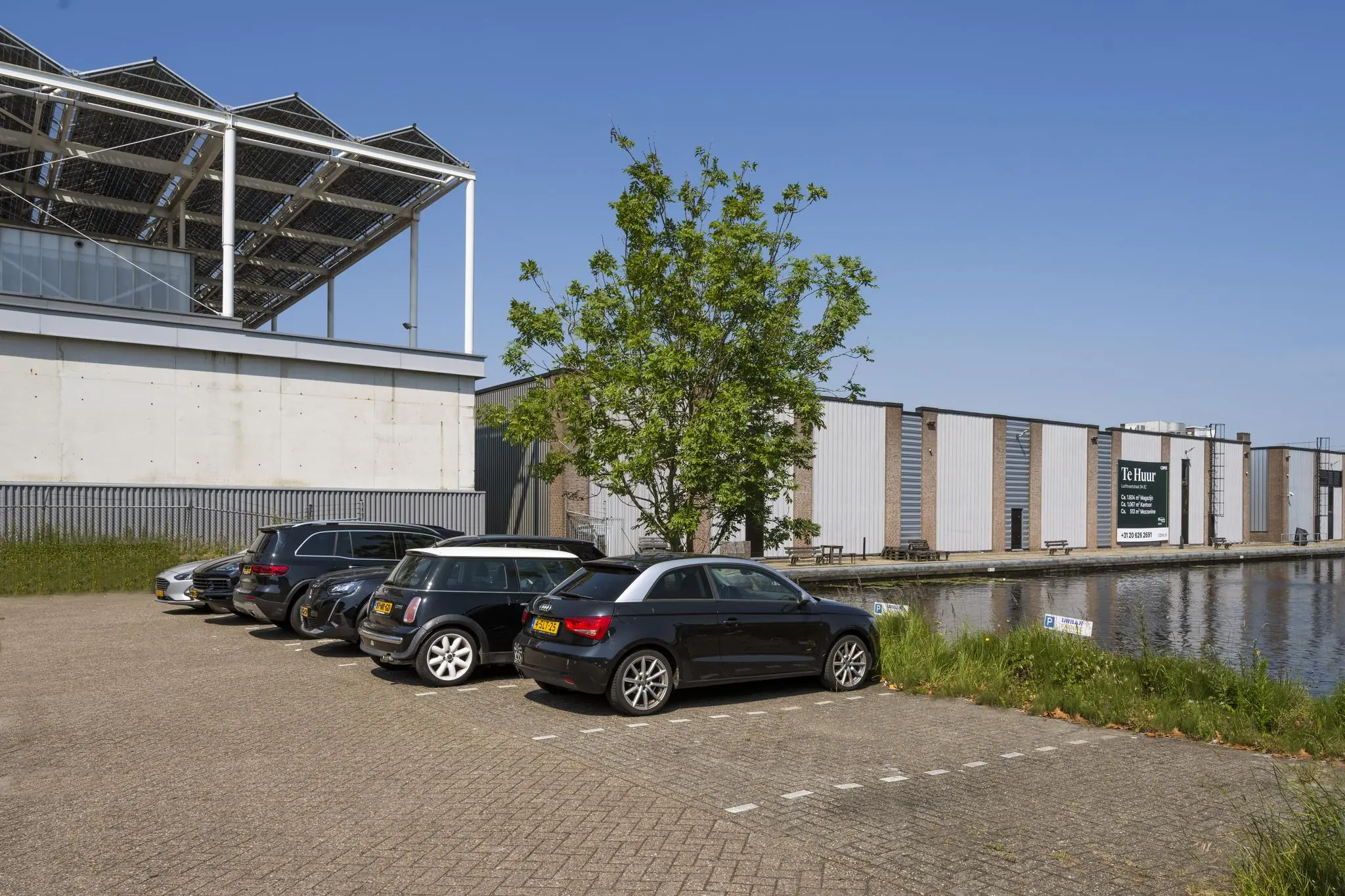 Parkeerplaats met geparkeerde auto's aan de Anthony Fokkerweg, naast een kanaal en bedrijfsgebouwen met een bord 'Te Huur'.