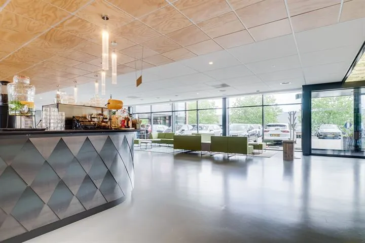 Moderne lobby met een koffiebar en groene zitbanken bij grote ramen met uitzicht op geparkeerde auto's.