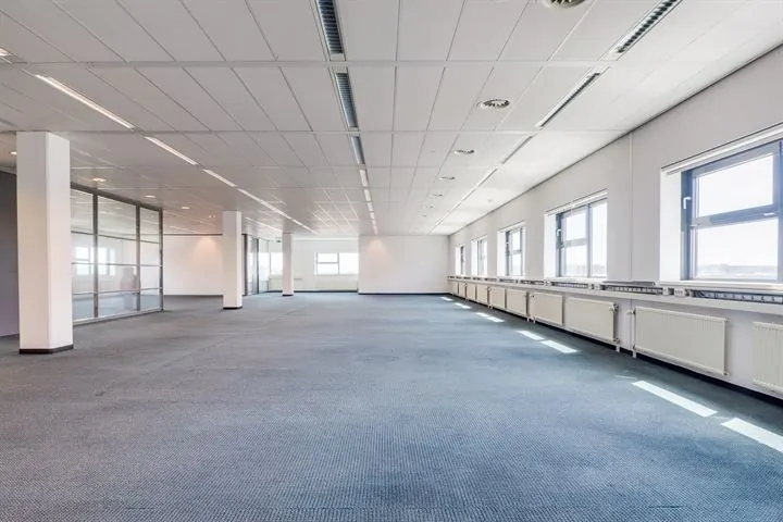 Lichte, lege kantoorruimte met blauw tapijt, systeemplafond en grote ramen aan de rechterkant in het gebouw aan de Ringwade.