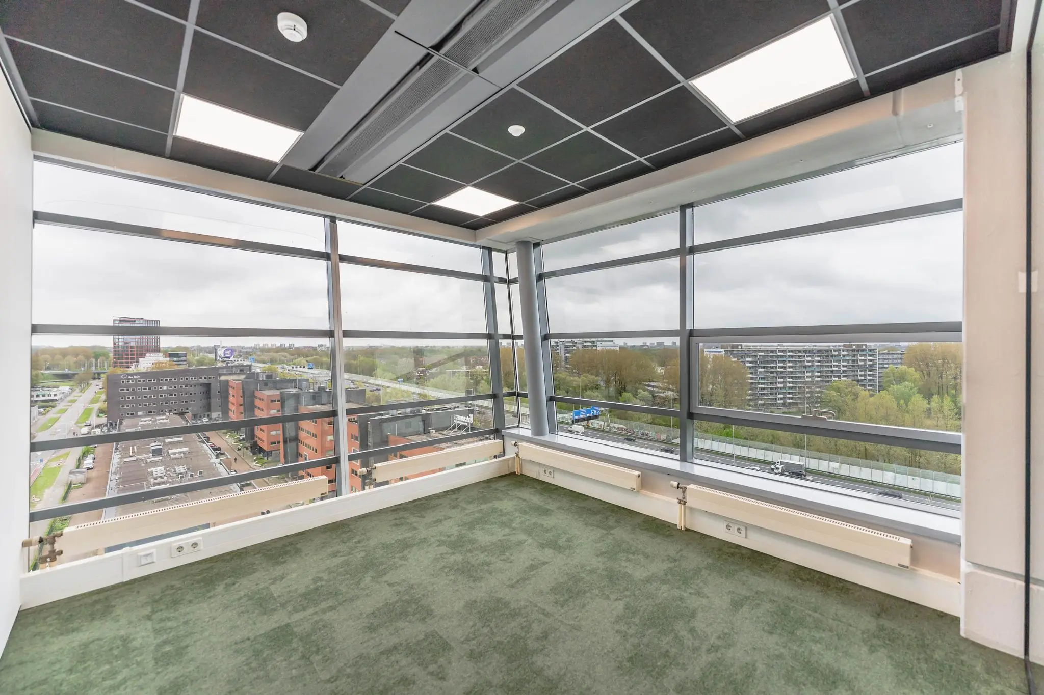 Leeg kantoor met groen tapijt en ramen van vloer tot plafond met uitzicht op Joop Geesinkweg in Amsterdam.