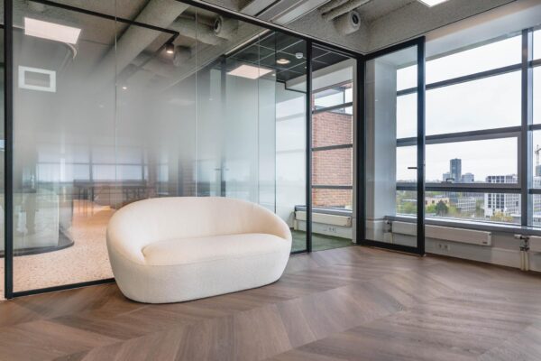 Moderne wachtruimte aan de Joop Geesinkweg met een crèmekleurige bank, glazen wanden en uitzicht op Amsterdamse kantoorgebouwen.