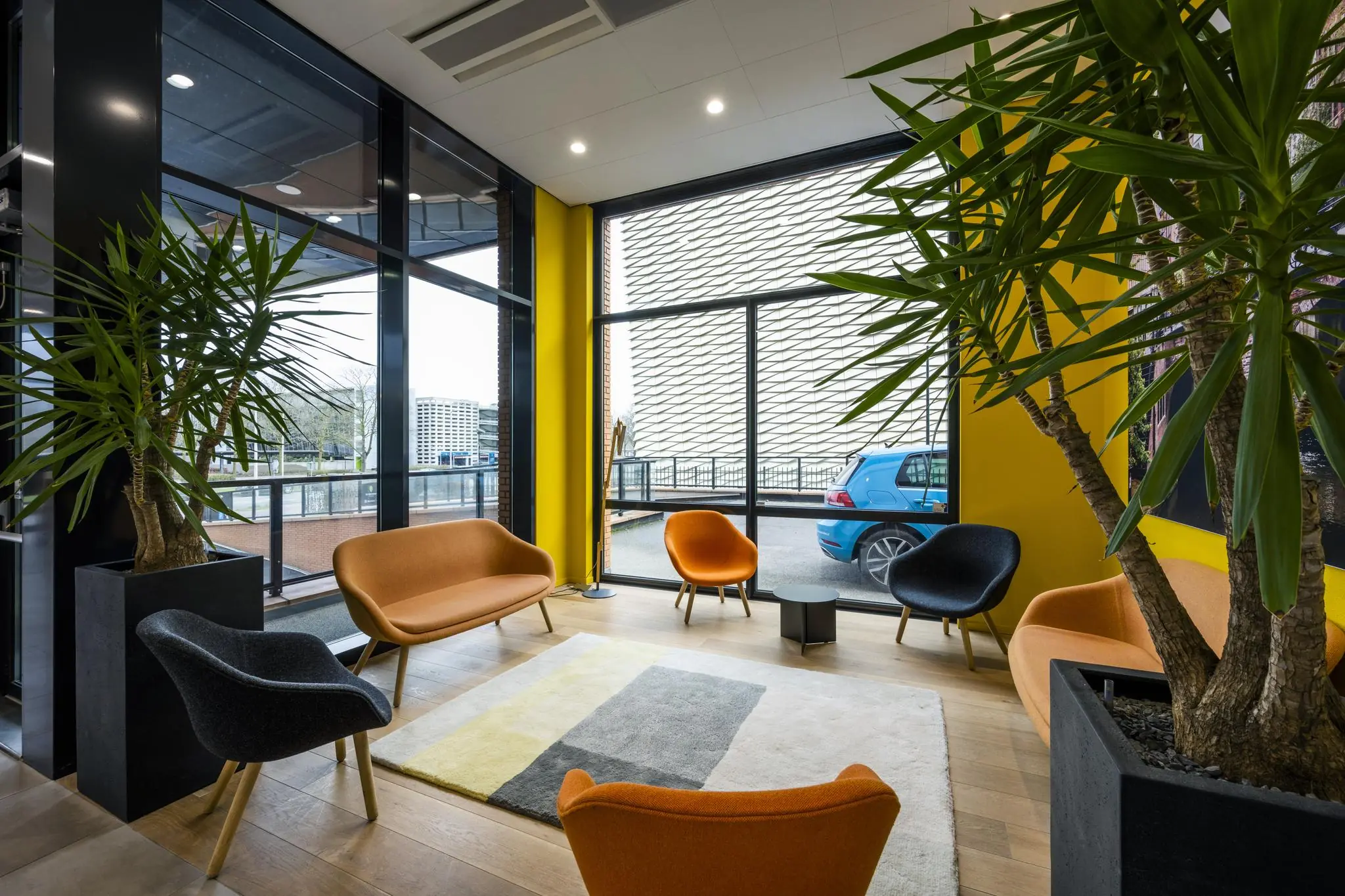 Moderne wachtruimte aan de Winthontlaan met kleurrijke stoelen, grote planten en uitzicht op een blauwe auto buiten.