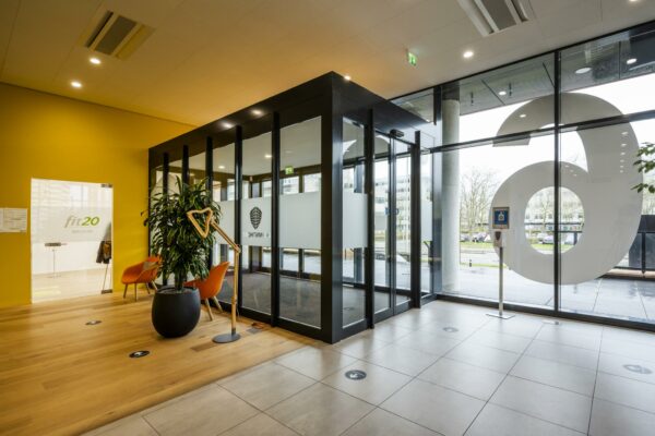 Moderne entree van een kantoorpand aan de Winthontlaan met zicht op de ingang van Fit20 en een wachtruimte met oranje stoelen.