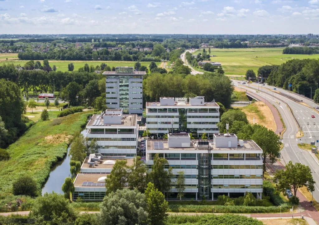 Luchtfoto van kantoorgebouwen aan de Burgemeester Stramanweg in een groen landschap met naastgelegen snelweg.