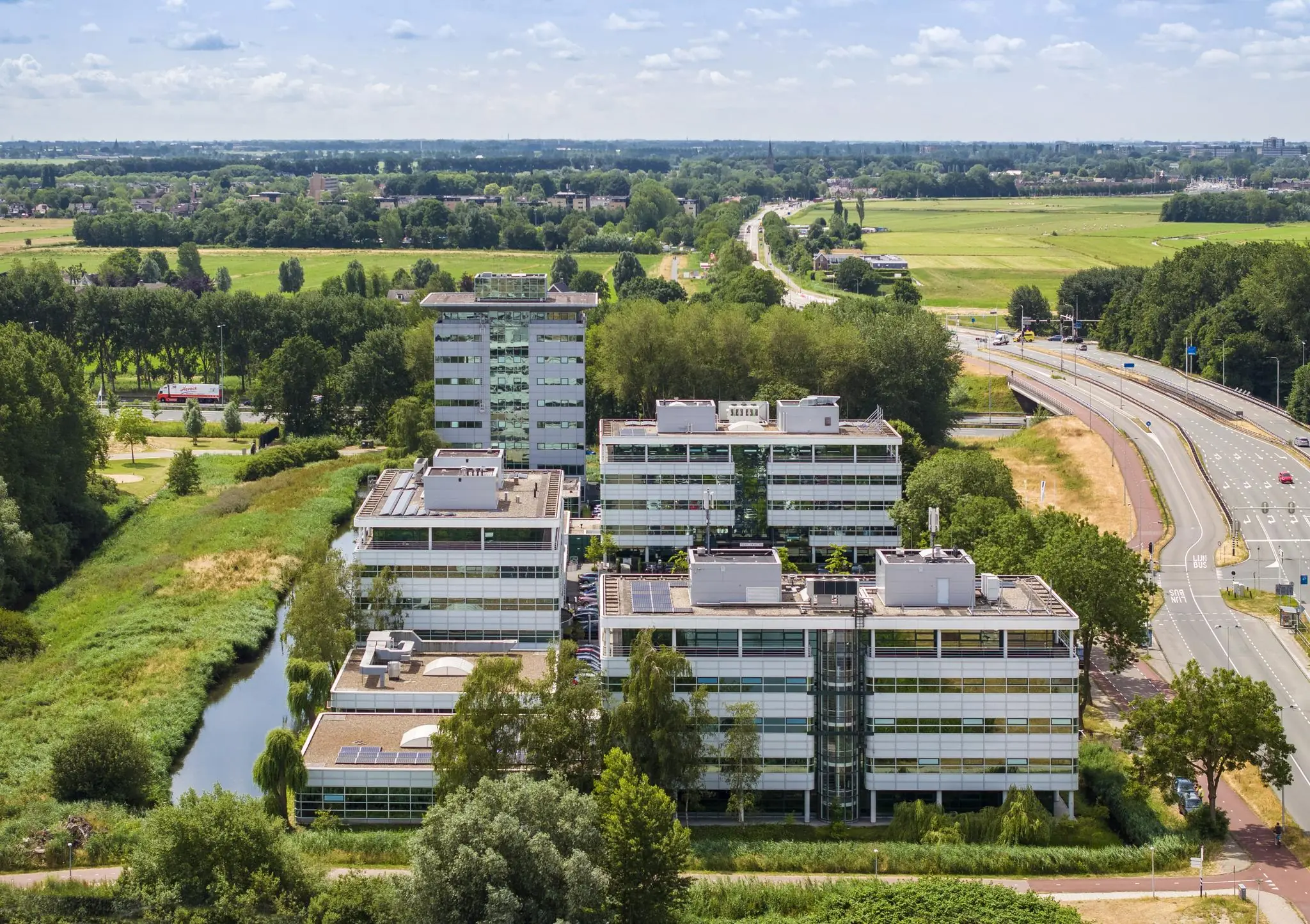 Luchtfoto van kantoorgebouwen aan de Burgemeester Stramanweg in een groen landschap met naastgelegen snelweg.