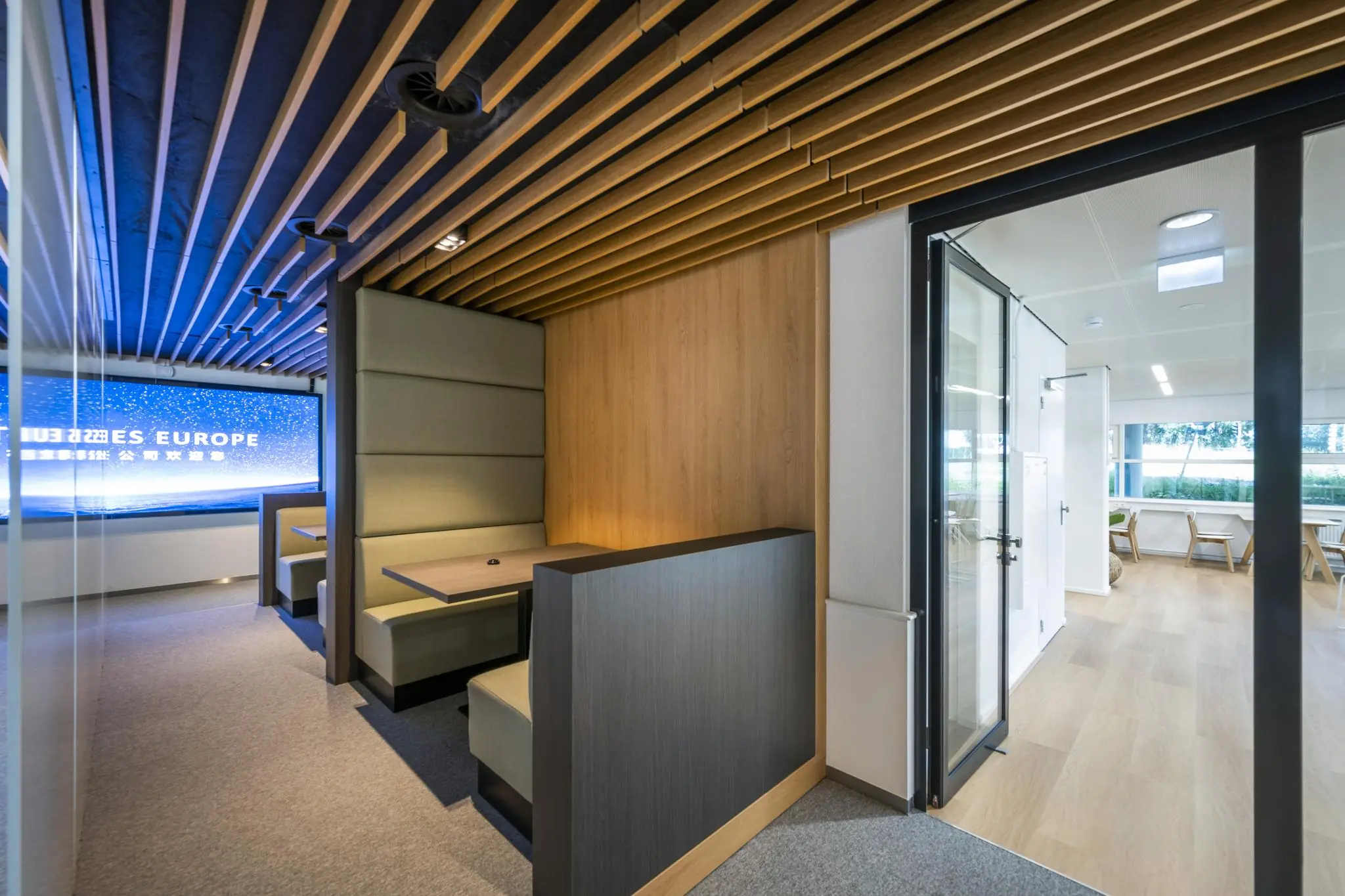 Moderne kantoorlounge aan de Burgemeester Stramanweg met houten plafondelementen, ingebouwde zitjes en een glazen deur naar een lichte vergaderruimte.
