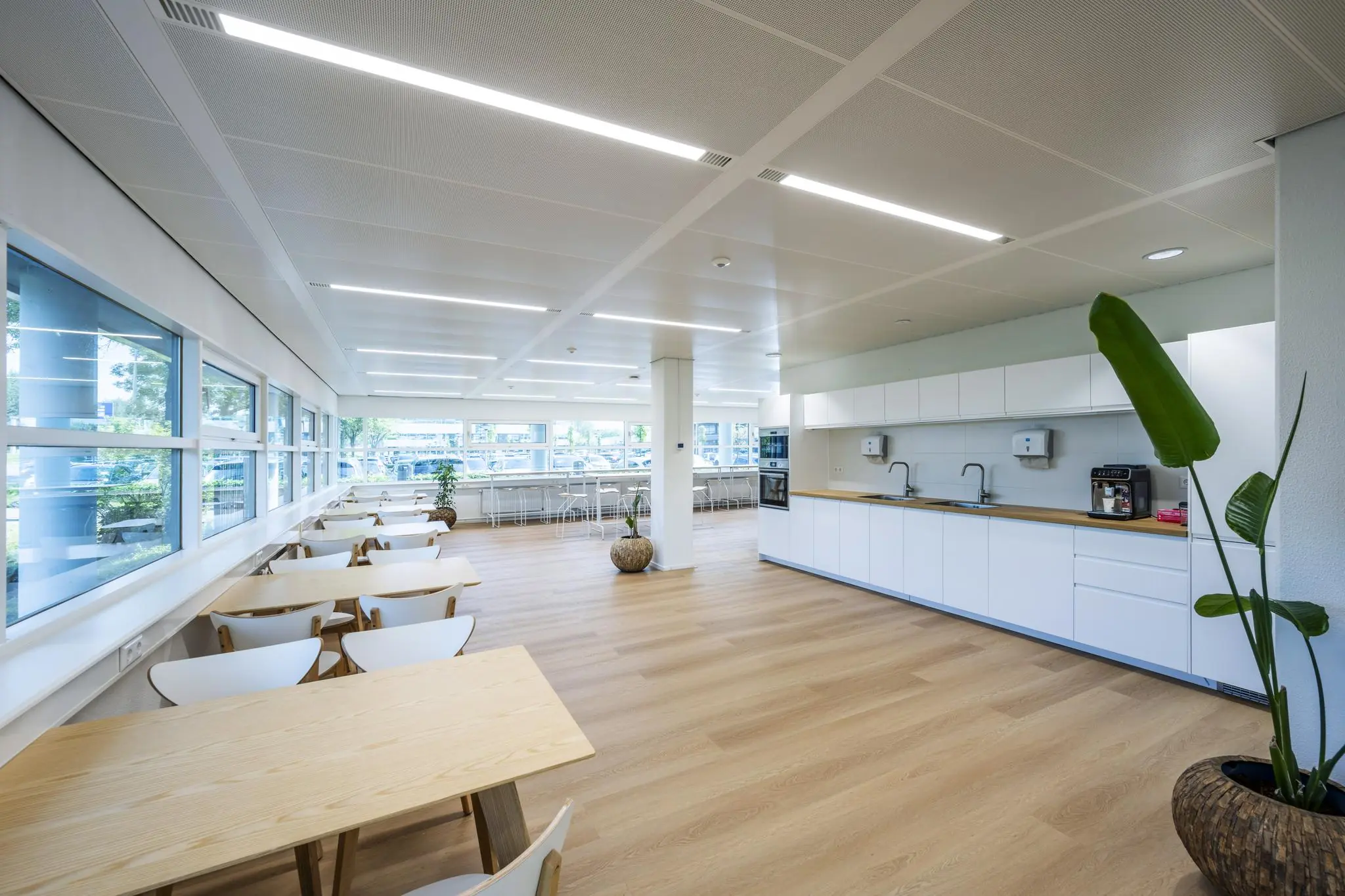 Moderne, lichte bedrijfskantine aan de Burgemeester Stramanweg met houten meubels, witte keuken en grote ramen.