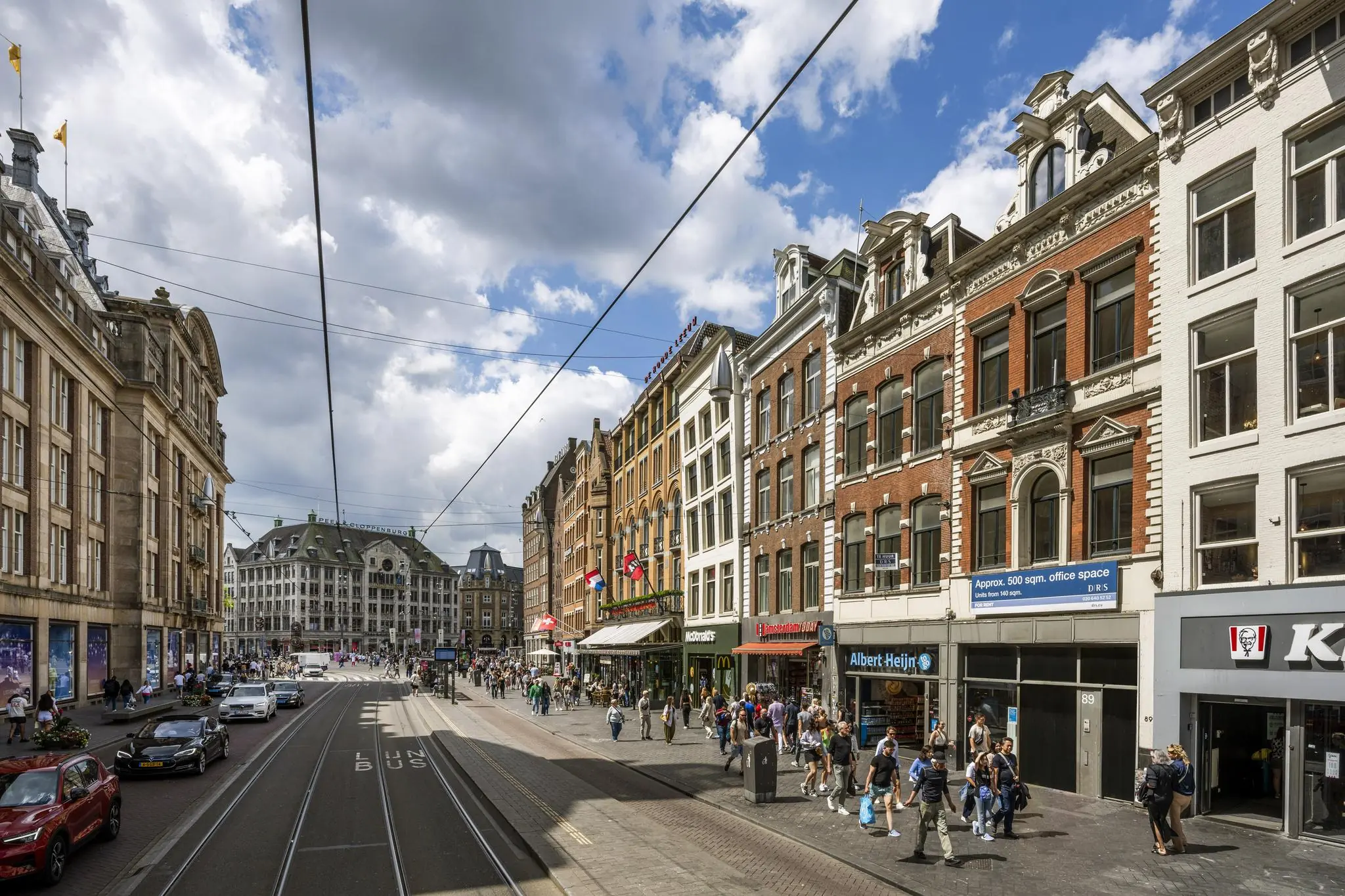 Drukke winkelstraat Damrak in Amsterdam met historische gevels, winkels en voorbijgangers op een zonnige dag.