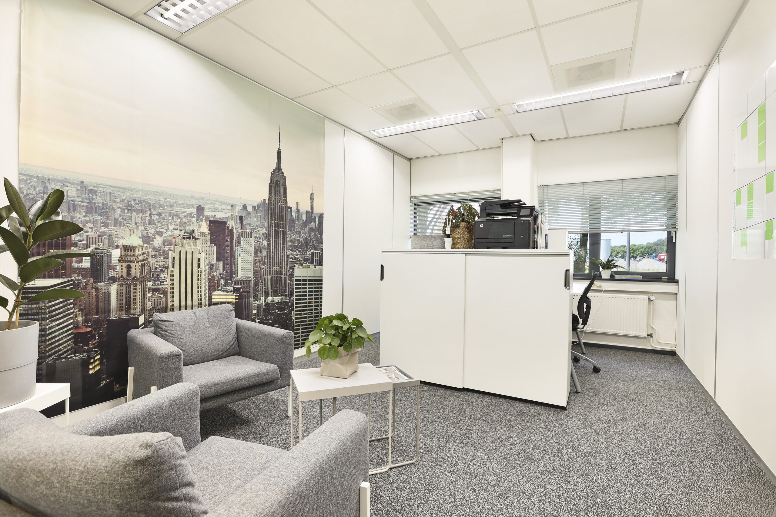 Kleine kantoorunit met grijze stoelen, planten en een groot fotobehang van een stadsgezicht.