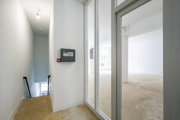 Lege commerciële ruimte aan het Damrak met zicht op een trap, brandmeldpaneel en grote ramen in een kale vloer.