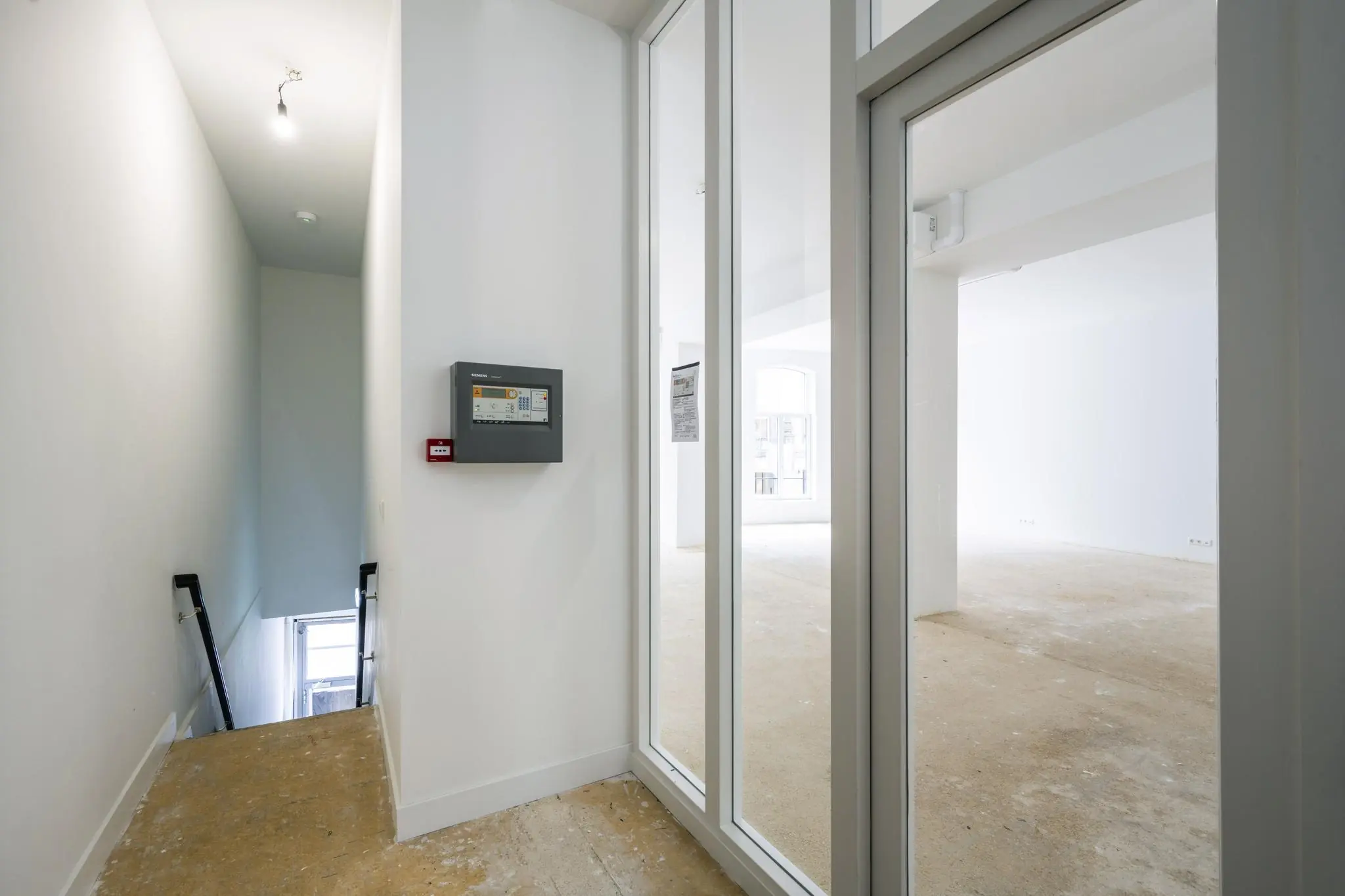 Lege commerciële ruimte aan het Damrak met zicht op een trap, brandmeldpaneel en grote ramen in een kale vloer.