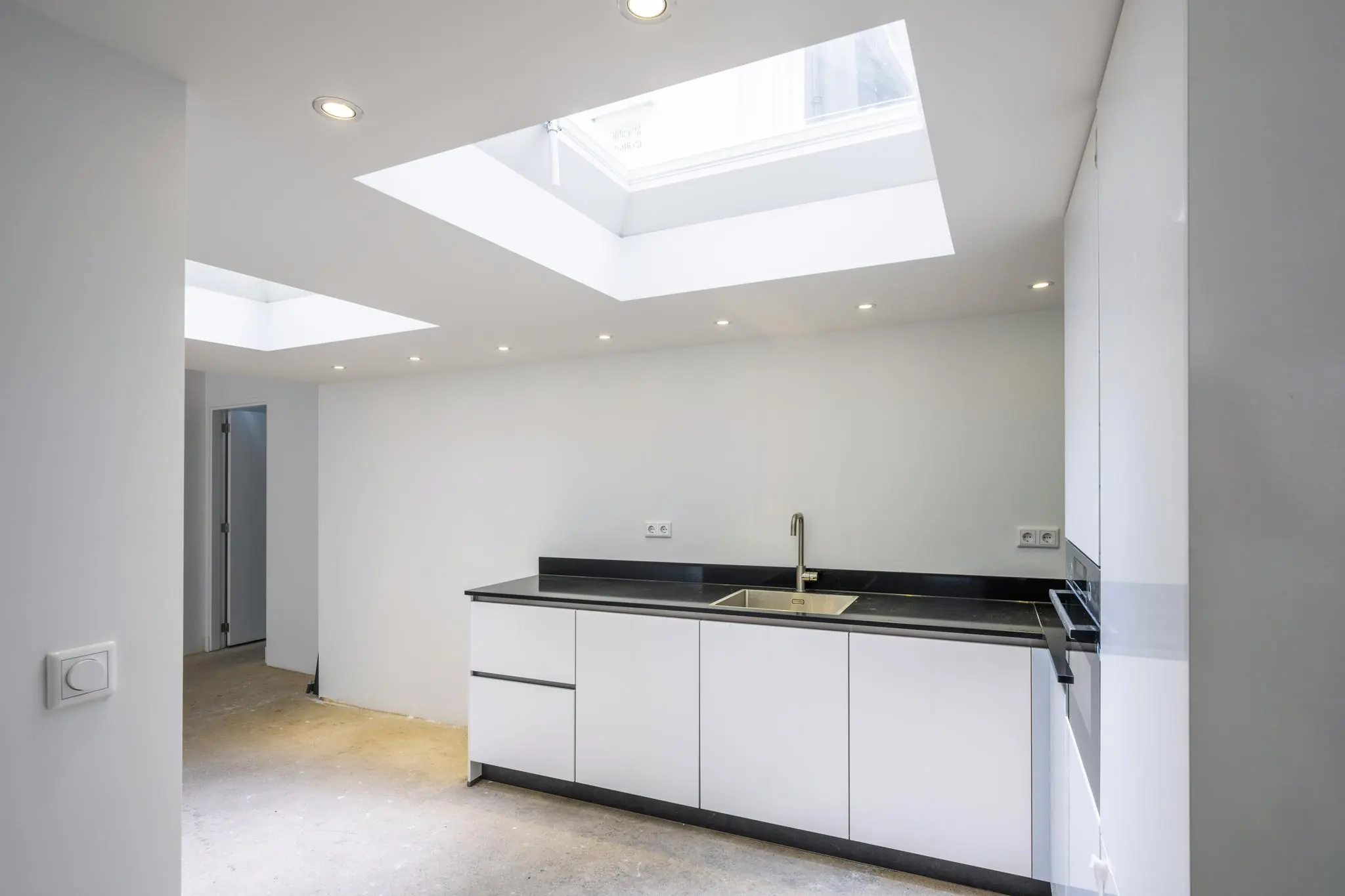 Moderne, minimalistische keuken met witte kasten, zwarte werkbladen en twee grote lichtkoepels in het plafond.