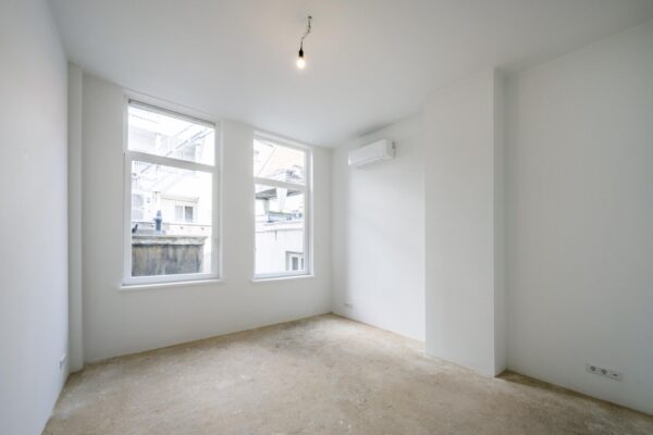 Leeg, licht appartement aan het Damrak met onafgewerkte vloer, witte muren en een airco-unit.