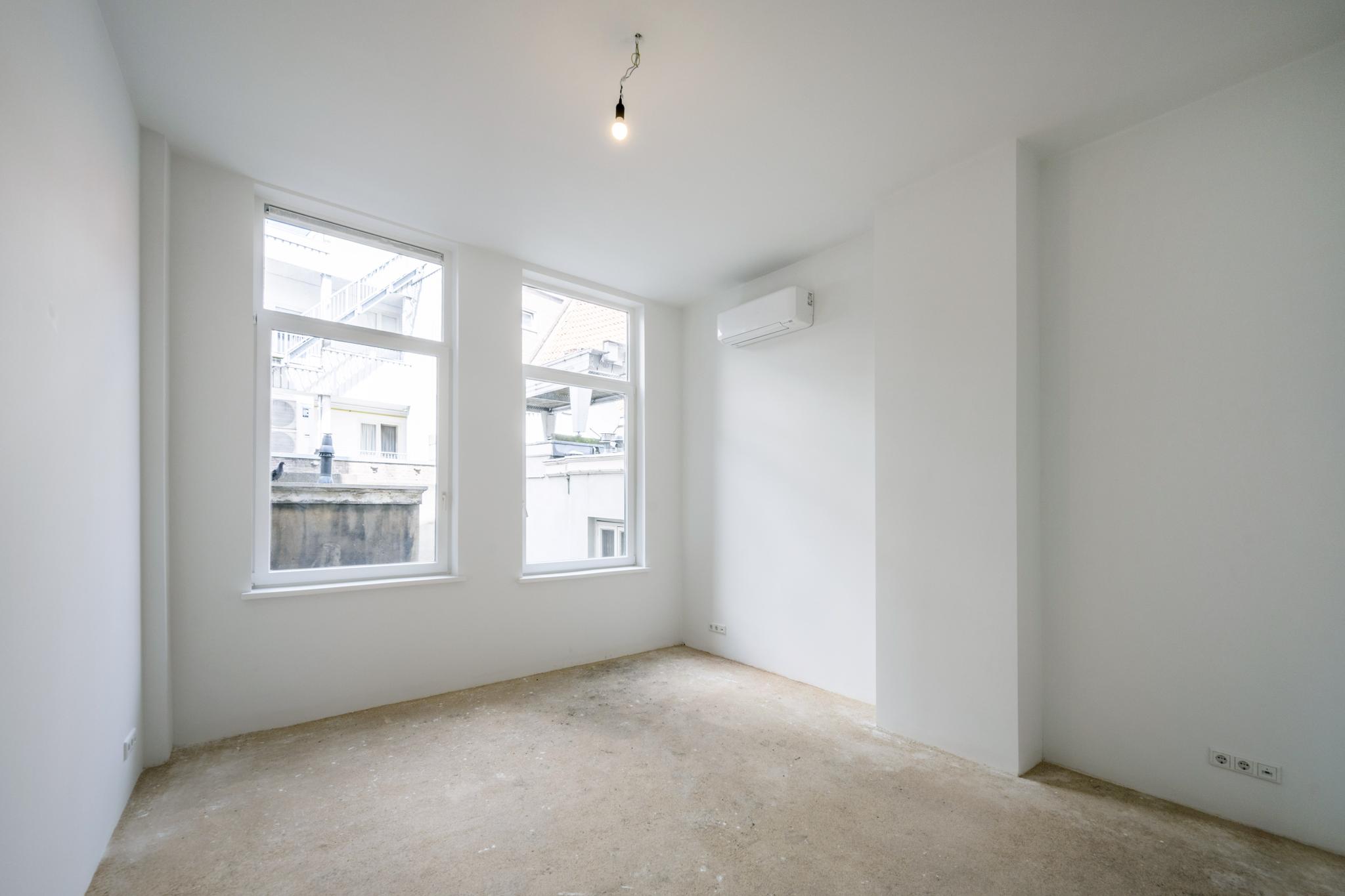 Leeg, licht appartement aan het Damrak met onafgewerkte vloer, witte muren en een airco-unit.