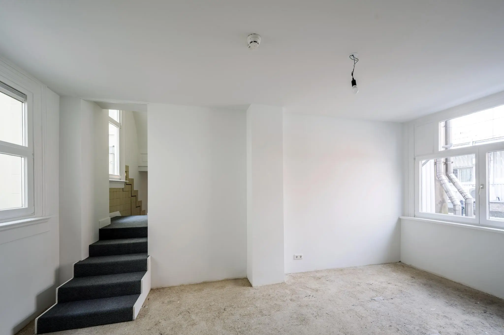 Lege kamer in een woning aan het Damrak met witte muren, ramen met zicht op een binnenplaats en een trap met donkere vloerbedekking.