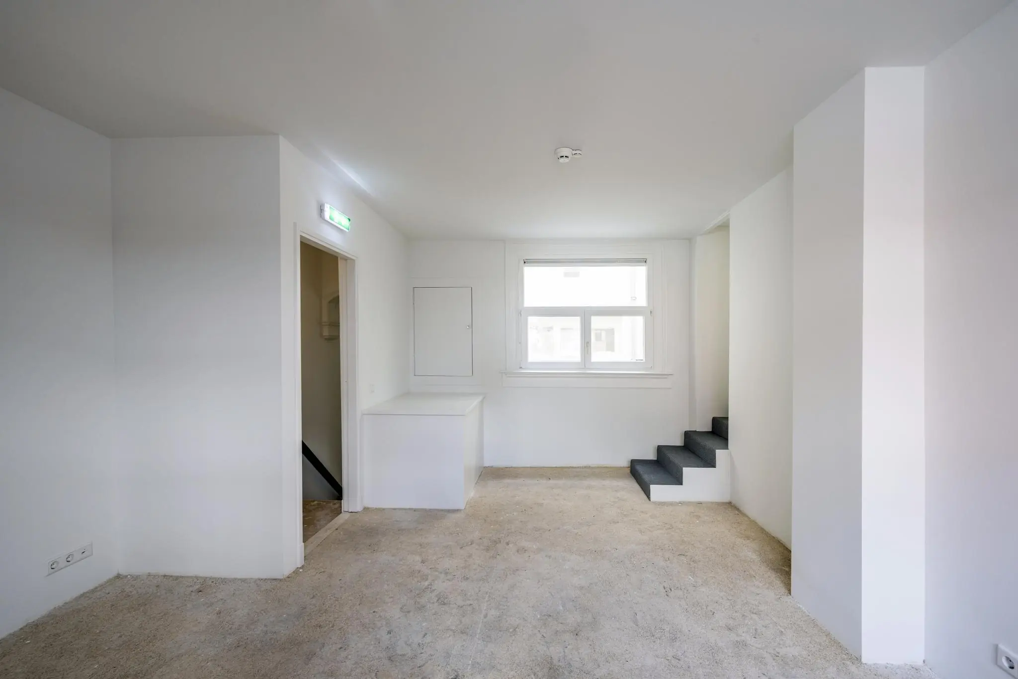 Lege, witte kamer aan het Damrak met betonnen vloer, raam en trapje naar een hoger gelegen ruimte.