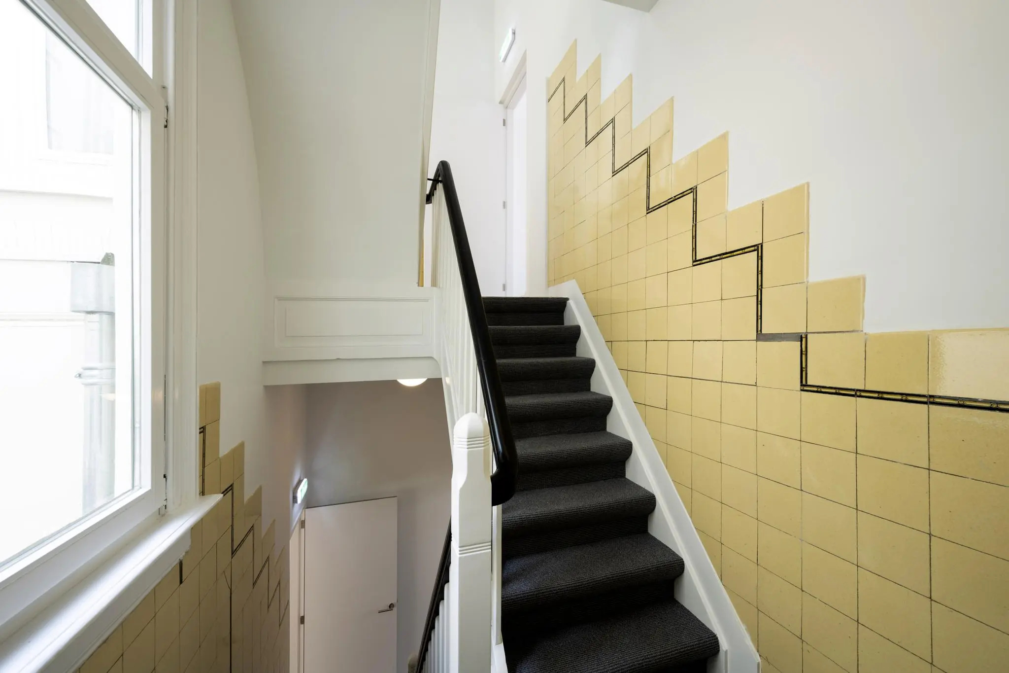 Trapopgang in een gebouw aan het Damrak met gele wandtegels en zwarte trapbekleding.