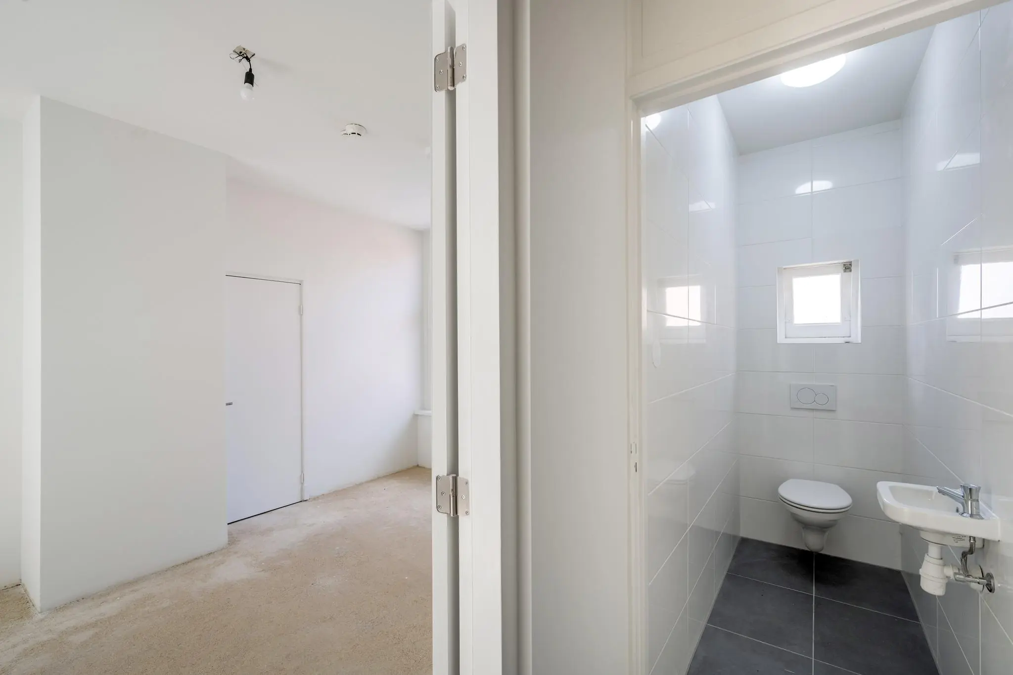 Wit betegeld toilet met kleine wastafel en raam, grenzend aan een lege, onafgewerkte kamer aan het Damrak.