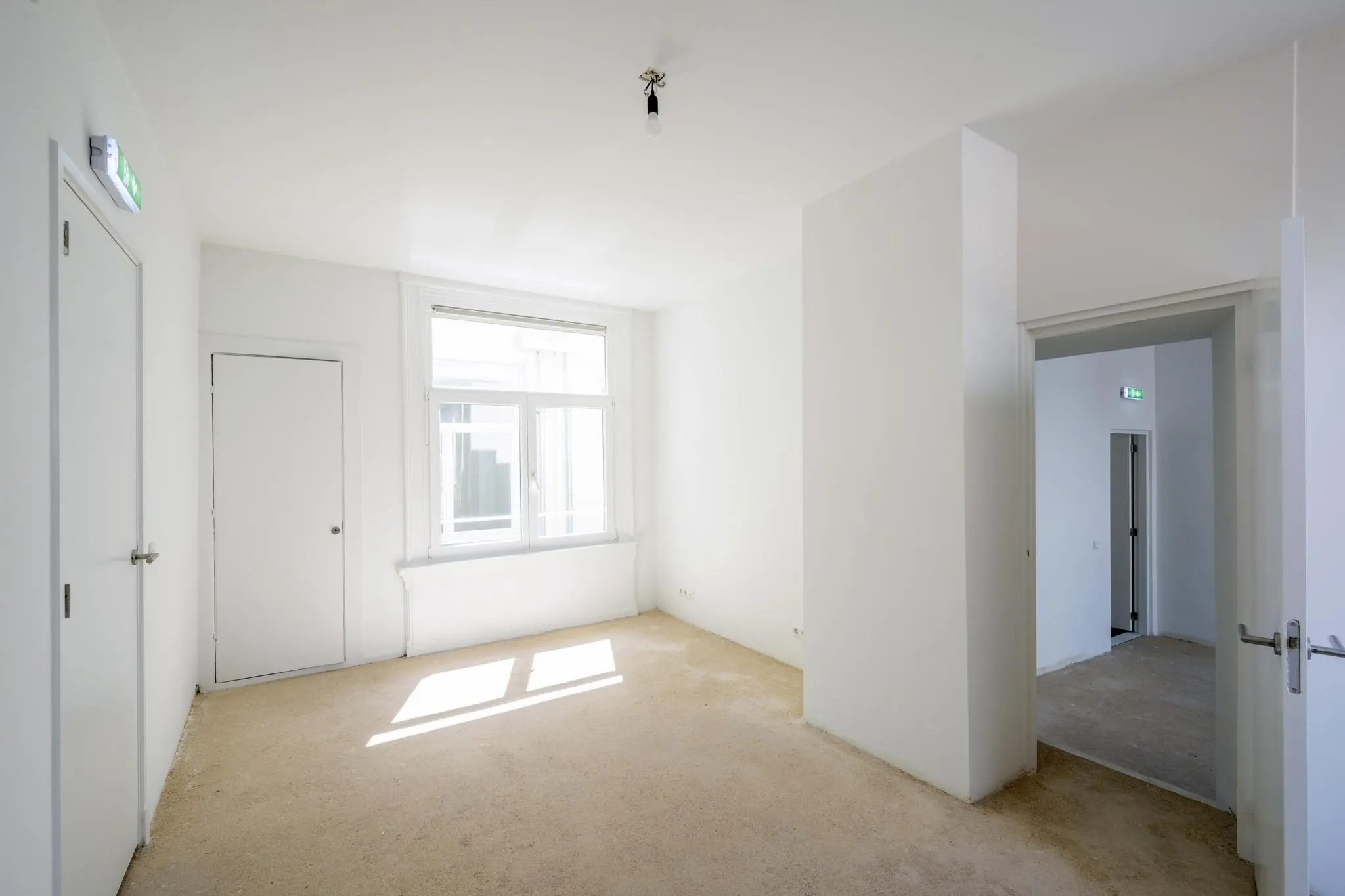 Lichte, lege kamer aan het Damrak met witte muren, een groot raam en onafgewerkte vloer.