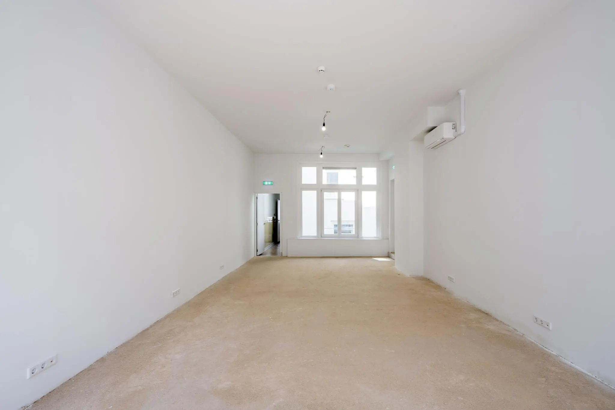 Leeg en witgeschilderd interieur van een ruimte aan het Damrak met grote ramen en een betonnen vloer.