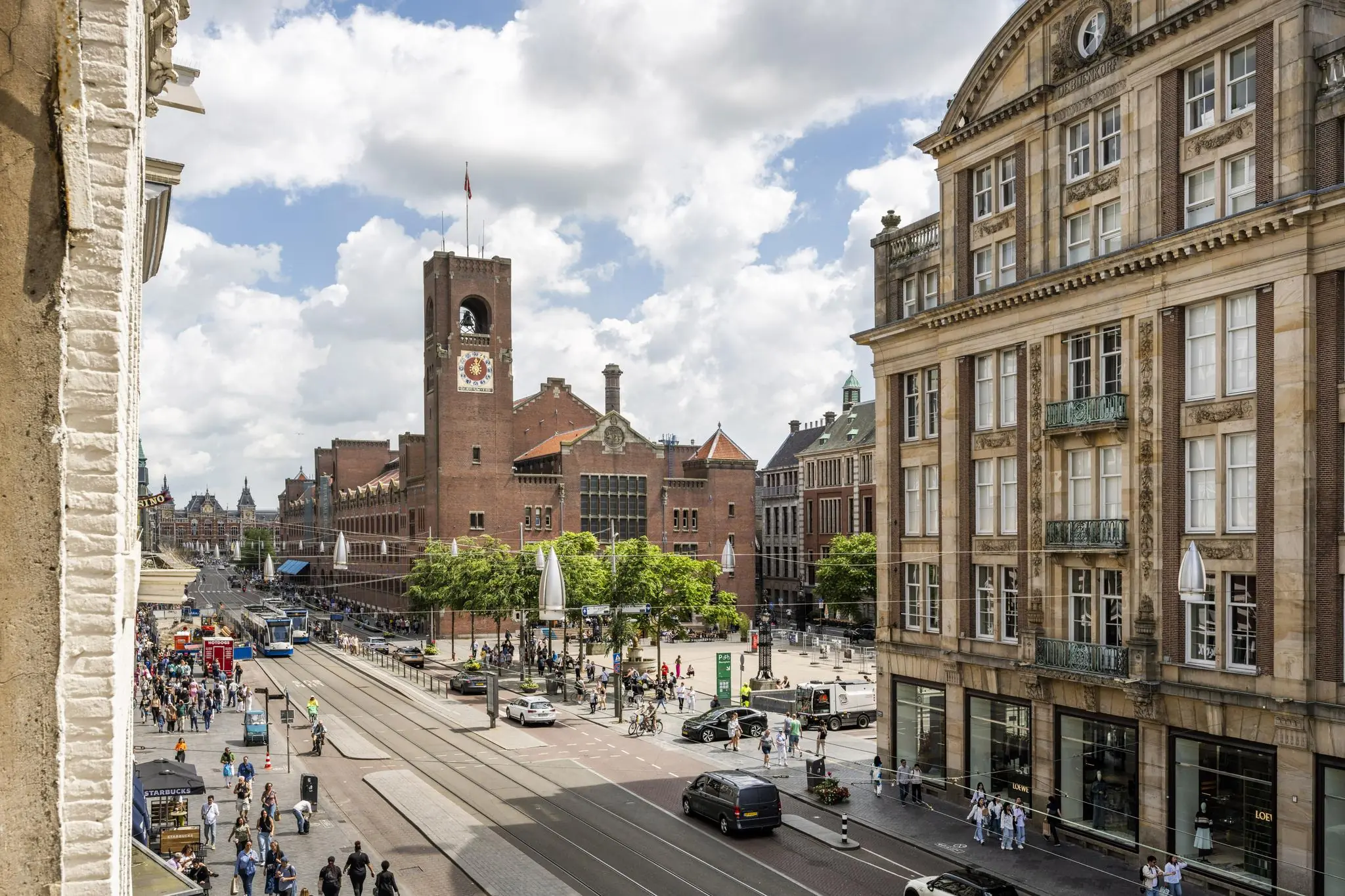 Drukke straat met trams, voetgangers en historische gebouwen aan het Damrak in Amsterdam, met de Beurs van Berlage op de achtergrond.