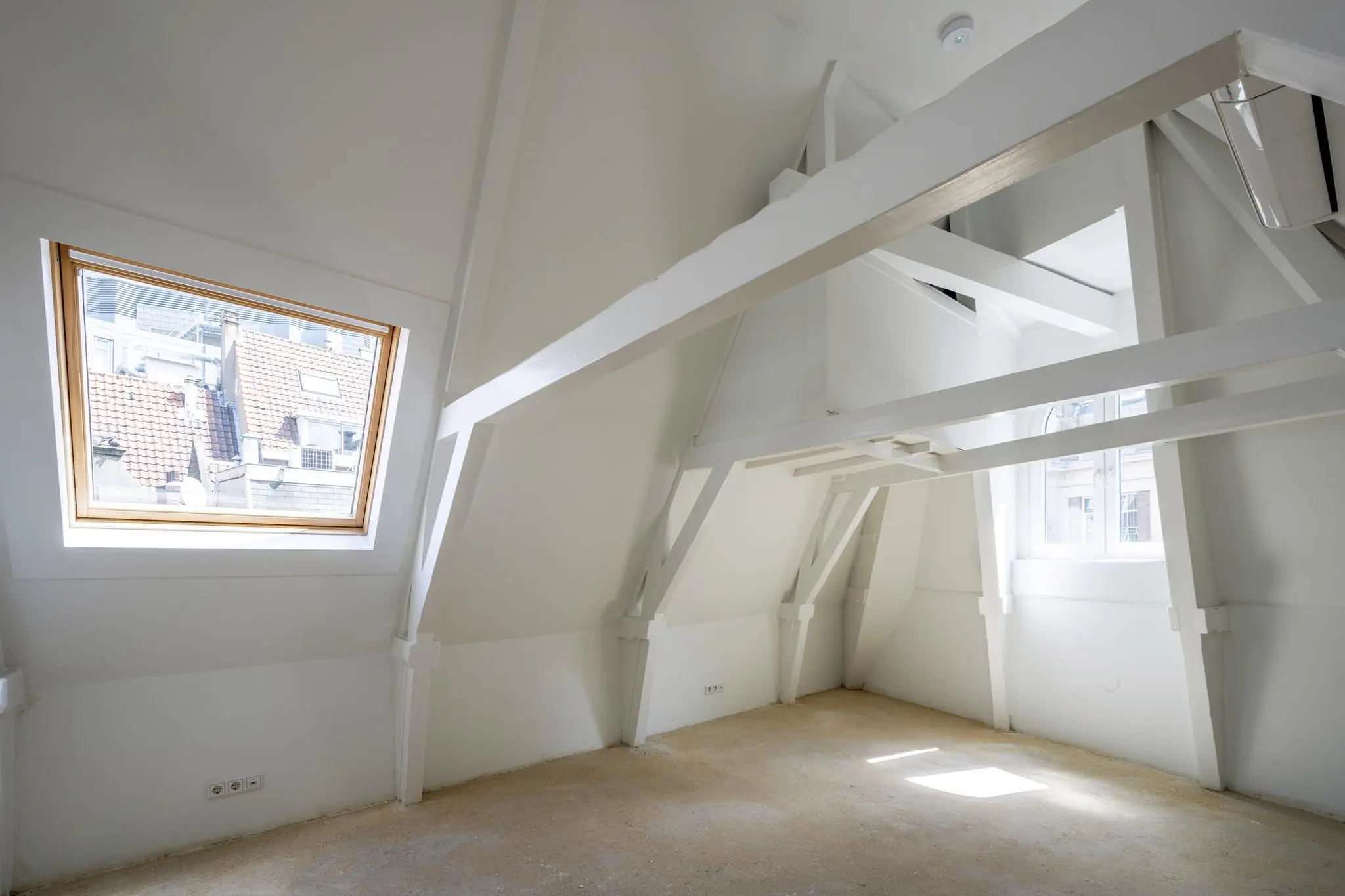 Zolderkamer aan het Damrak met witte houten balken, schuine wanden en twee ramen die uitzicht bieden op omliggende gebouwen.