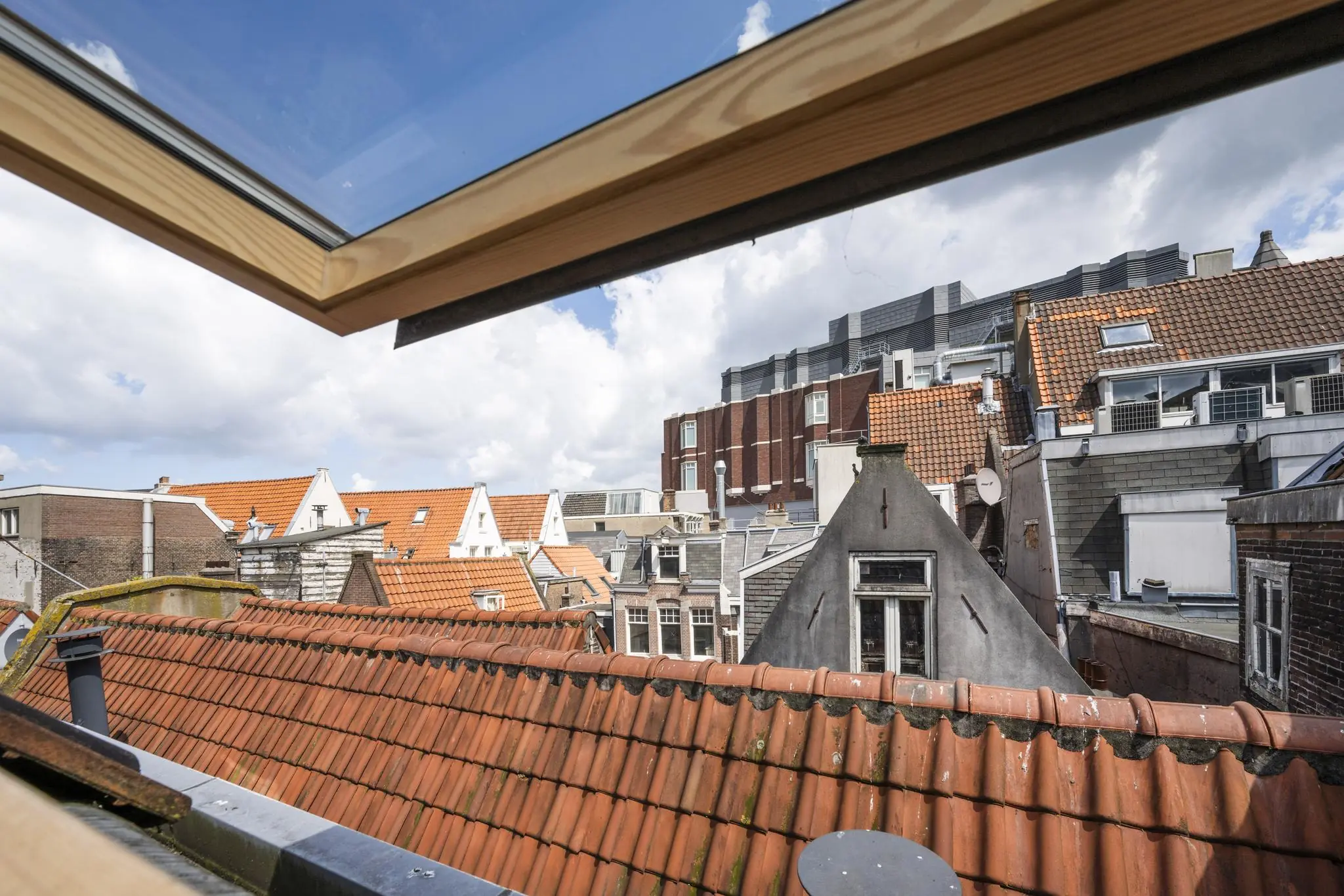 Uitzicht vanuit een open dakraam op de rood/oranje pannendaken en gevels van oude gebouwen aan het Damrak in Amsterdam.